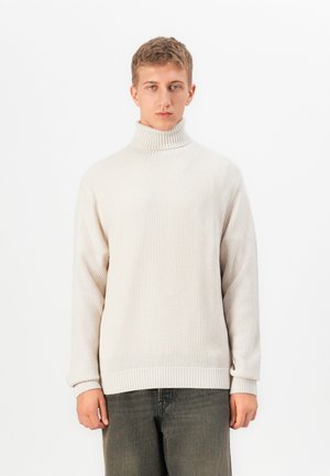 Jonge man in een crèmekleurige coltrui en donkergrijze pantalon, staande tegen een effen witte achtergrond.