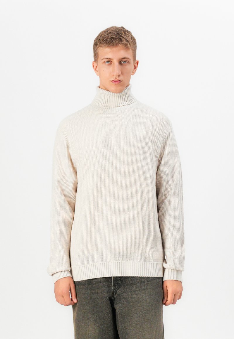 Young man wearing a cream turtleneck sweater and dark gray pants, standing against a plain white background.