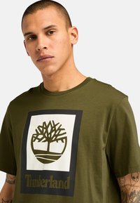 Olivově zelené bavlněné tričko s bílým čtvercovým logem a designem stromu, pod kterým je v tmavě olivové barvě text "Timberland". Krátké rukávy.