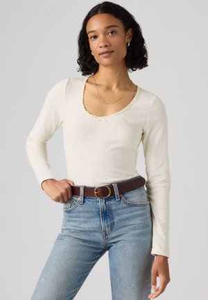 Femme aux cheveux bouclés et foncés porte un haut à manches longues crème avec un col en arrondi festonné, un jean bleu taille haute et une ceinture marron, debout, détendue.