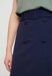 Tmavě modrá sukně s texturovaným povrchem, vybavená čtvercovým páskem a ozdobnými knoflíky na přední straně, představující prvky propracovaného střihu.