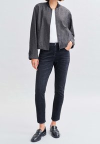 Chaqueta bomber gris con tejido texturizado, cuello negro, llevada sobre una camisa blanca; combinada con jeans oscuros y zapatos negros sin cordones.