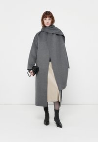 Grijze wollen jas met een open voorkant, oversized ontwerp en brede mouwen. Gecombineerd met een beige rok en zwarte accessoires. Gladde textuur.