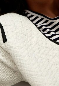 Tissu blanc texturé présentant un motif tissé, accompagné d'un col rayé noir et blanc. Les détails incluent une bordure sombre le long de la manche.