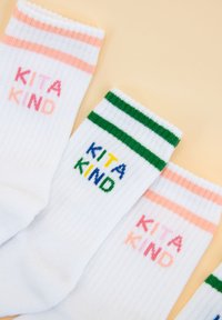 Białe prążkowane skarpetki mają kontrastowe poziome paski w kolorze różowym i zielonym na górze. Fraza "KITA KIND" jest wyhaftowana w różnych kolorach.