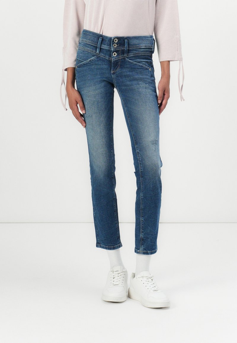 Blå denimjeans med en stram design, to-knapp liv, subtil fading og slitte detaljer. Kombinert med hvite sneakers og hvite sokker.