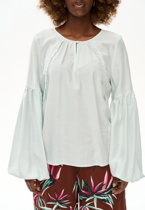 Kobieta ubrana w jasnoniebieską bluzkę z bufkami i brązowe spodnie w kwiatowy wzór, pokazana od szyi do bioder.