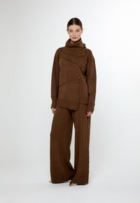 Brun stickad tröja med hög krage och sidodetaljer, i kombination med vida byxor med dekorativa knappar längs sidorna.