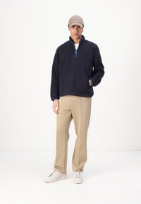 Navy kék fleece pulóver fél cipzárral, kontrasztos kék díszítéssel és elülső zsebekkel, bézs nadrággal és fehér sportcipőkkel párosítva.