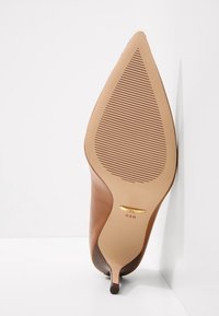 Brun läderklack med spetsig tå. Slät yta, smal klack och en ljusbrun gummisula med vertikala spår. Guldmärke synligt.