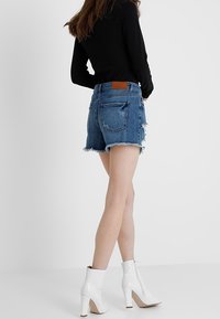 Žena v roztrhaných denimových šortkách, černém dlouhém rukávu a bílých vysokých botách na podpatku stojící na bílém pozadí.