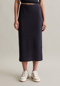 Marineblaue, strukturierte Midi-Rock mit einem taillierten Bund, kombiniert mit weißen und beigen Sneakers, auf einem neutralen Hintergrund stehend.