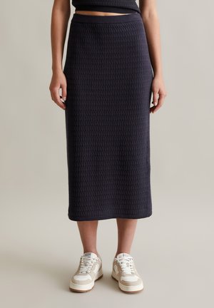 Jupe midi structurée bleu marine, avec une taille ajustée, associée à des baskets blanches et beiges, se tenant sur un fond neutre.