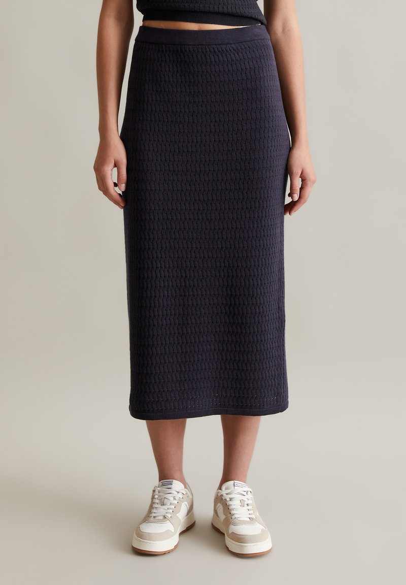 Marineblaue, strukturierte Midi-Rock mit einem taillierten Bund, kombiniert mit weißen und beigen Sneakers, auf einem neutralen Hintergrund stehend.