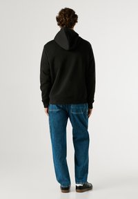 Μαύρη ζακέτα με κουκούλα, με θέα από πίσω, συνδυασμένη με μπλε φαρδύ τζιν και μαύρα παπούτσια σε ουδέτερο φόντο.