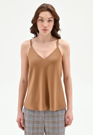 Jeune femme aux cheveux bruns mi-longs portant un haut sans manches marron à col en V et un pantalon à carreaux, debout devant un fond blanc.