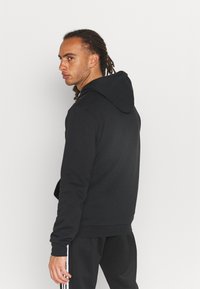 Sudadera negra de tejido suave con un bolsillo canguro. Cuenta con capucha ajustable y puños de canalé. Combinada con pantalones negros a juego.