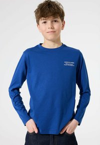 Junge, der mit Händen in den Taschen steht, trägt ein blaues Langarmshirt mit weißem Text auf der Brust vor einem schlichten weißen Hintergrund.