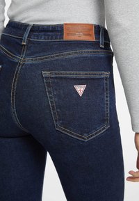Los jeans de mezclilla azul oscuro presentan una cintura alta, costuras visibles y un parche de logo triangular cerca del bolsillo trasero. Textura suave en general.