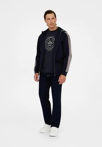 Hanorac bleumarin cu fermoar, accente gri, tricou cu guler rotund și logo circular, și pantaloni bleumarin asortați; asortate cu adidași albi.