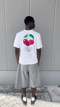T-shirt branca de manga curta com um gráfico colorido de cerejas nas costas, combinada com calções cinzentos até ao joelho e sapatos pretos.