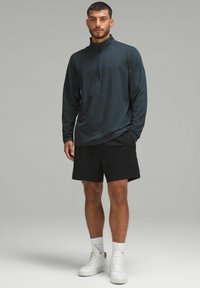 Pulover lung cu mânecă lungă de culoare teal închis, cu guler cu fermoar pe un sfert, asortat cu pantaloni scurți negri și adidași albi. Material moale și croială lejeră.