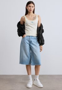 Φωτεινό κίτρινο ριμπ τοπ με μπλε λογότυπο, μαύρο μπουφάν bomber, χαλαρές ανοιχτό μπλε τζιν κολότ, λευκά αθλητικά παπούτσια και λευκές κάλτσες αστραγάλου.