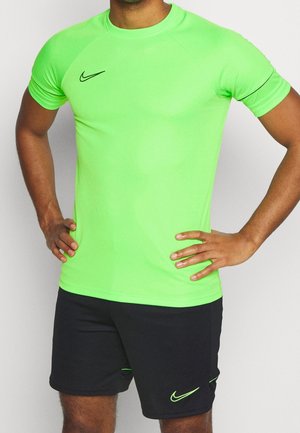 Homme portant un t-shirt de sport Nike manches courtes vert vif et un short Nike noir, debout les mains sur les hanches devant un fond uni.
