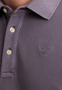 Ljubičasta polo majica s teksturiranim materijalom, tri gumba i izvezeni logotip s orlom na prsima.
