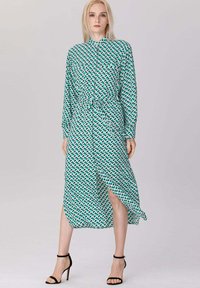 Femme blonde portant une robe midi ceinturée à manches longues avec un motif géométrique turquoise et blanc, assortie de sandales à talons hauts noires.