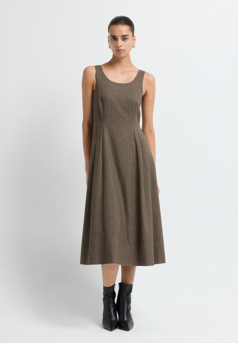 Vestido sin mangas de cuadro marrón con silueta en A, que presenta un cuerpo ajustado y una falda fluida. Se lleva con botas negras hasta el tobillo.