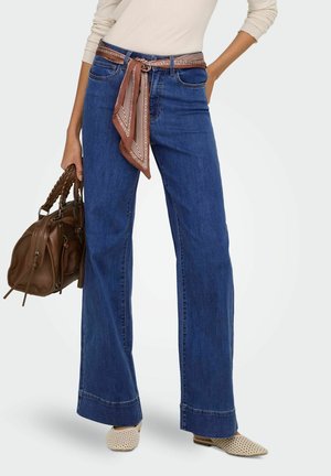 Femme portant un jean bleu à jambes larges avec une ceinture écharpe marron à motifs, un haut beige à manches longues, tenant un sac à main marron et portant des chaussures tissées beiges.