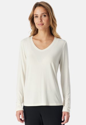Femme aux cheveux châtain clair portant un haut blanc à manches longues avec col en V et un pantalon noir, debout devant un fond uni.