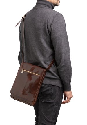 Man draagt een donkergrijze trui en spijkerbroek en draagt een glanzende bruine leren crossbodytas met een ritsvak aan de voorkant.