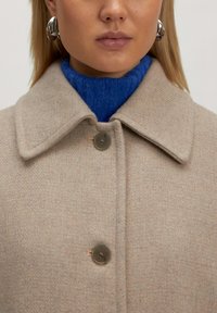 Beige Wollmantel mit breitem Kragen, ausgestattet mit einem Knopfverschluss und einer strukturierten Oberfläche. Darunter ist ein leuchtend blauer Rollkragenpullover sichtbar.
