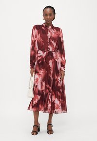Langes, gefältetes Kleid mit rotem und pinkem abstraktem Muster, geknöpftem Kragen, langen Ärmeln und tailliertem Gürtel. Kombiniert mit einer weißen Handtasche und schwarzen Sandalen.