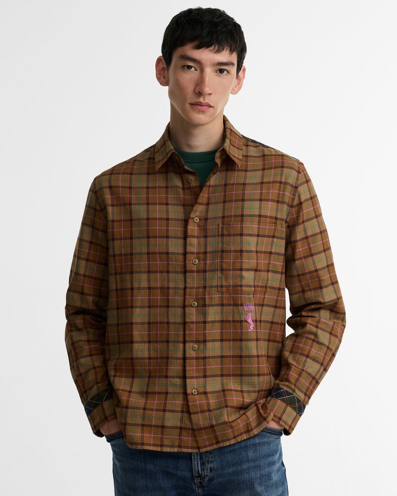Jeune homme portant une chemise à carreaux marron et verte à boutons avec un motif brodé rose sur la poche et un jean bleu.