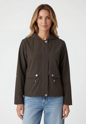 Vrouw met een donkerbruine jas met capuchon en drukknoopsluiting, en lichtblauwe jeans, staand tegen een effen witte achtergrond.