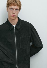 Schwarze Wildlederjacke mit einem Reißverschluss vorne, Punktkragen und einer Brusttasche. Die glatte Textur und das moderne Design unterstreichen ihren minimalistischen Look.