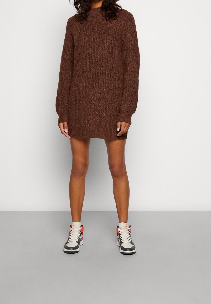 Personne portant une robe-pull à manches longues en tricot marron et des baskets montantes blanches, noires et rouges, debout contre un mur blanc uni.