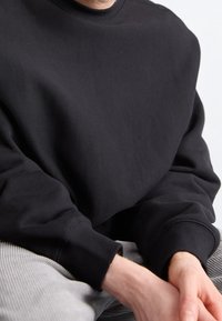 Persoon in een zwart sweatshirt met lange mouwen en lichtgrijze broek, met handen op hun schoot tegen een effen achtergrond.