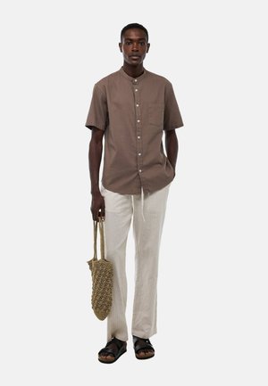 Homme portant une chemise marron à manches courtes boutonnée, un pantalon beige clair, des sandales noires, tenant un petit sac en tissu tissé dans sa main droite.