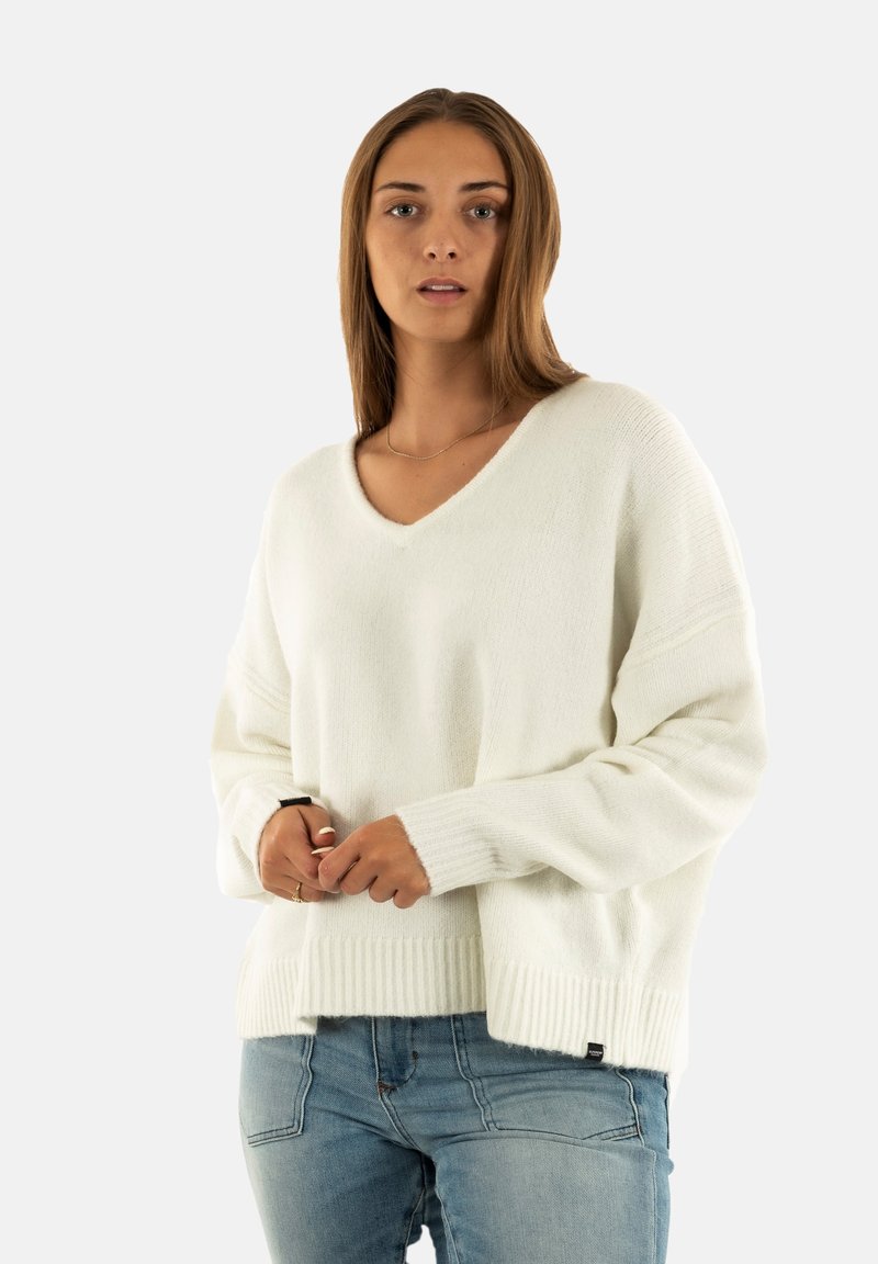 Giovane donna con capelli castani lisci, che indossa un maglione bianco a scollo a V e jeans blu, in piedi davanti a uno sfondo chiaro e uniforme.