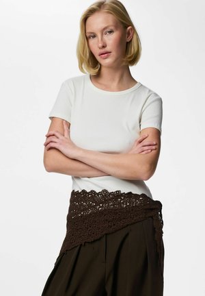 Femeie blondă cu păr scurt, purtând un top alb cu mâneci scurte și pantaloni închiși la culoare, cu o centură maronie croșetată, cu brațele încrucișate, pe un fundal neutru.