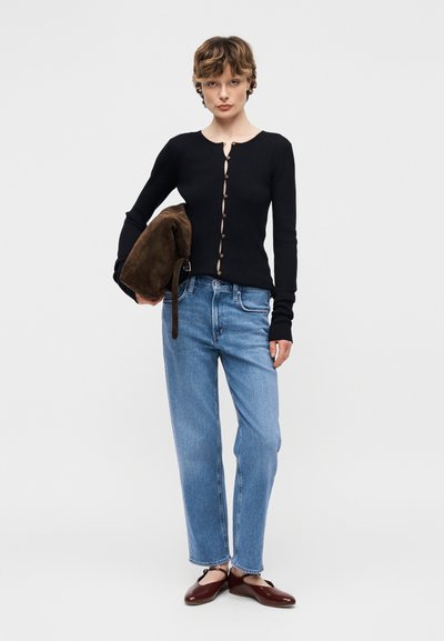 Γυναίκα που στέκεται φορώντας μπλε τζιν, μαύρο πουλόβερ με κουμπιά, κρατώντας καφέ γούνινο μπουφάν, και φοράει σκούρα κόκκινα παπούτσια Mary Jane μπροστά σε απλή φόντο.