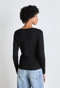 Tricou negru cu mânecă lungă, cu un model texturat, având un guler larg și o siluetă ajustată, asortat cu blugi lejeri albaștri deschiși.