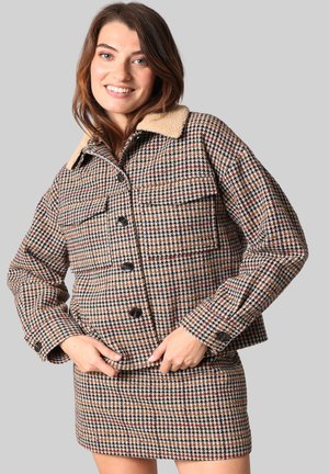 Veste à motif pied-de-poule avec un col beige, dotée de boutons noirs et de deux poches avant. Le tissu a une apparence texturée et tissée.
