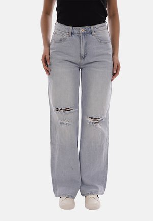 Donna che indossa jeans larghi a vita alta strappati di colore grigio chiaro con sneakers bianche e una canotta nera, in piedi dritta contro uno sfondo bianco.