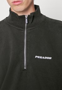 Černá poloviční zipová fleecová mikina s žebrovaným límcem. Na levé straně má bílé logo "PEGADOR". Měkká textura, volný střih.