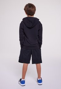 Černá mikina s kapucí a klokaní kapsou, doplněná černými síťovanými šortkami. Outfit je dokončen bílými a modrými teniskami.